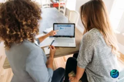 Two women reviewing financial data on a laptop, discussing financial education strategies.