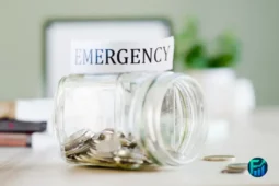 Glass jar labeled “Emergency” partially filled with coins, symbolizing saving for financial emergencies.