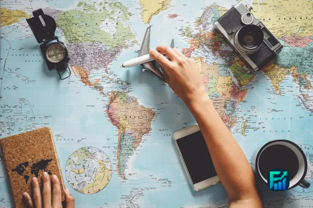 Paying for trips: hands hold a credit card and small plane over a world map with a camera, compass, phone and coffee nearby