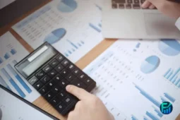 Person using a calculator on financial reports and charts, reviewing expenses and managing money to avoid Overdraft Fees.
