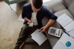 Man sitting on the floor reviewing documents with a calculator and phone, concerned about medical bills.