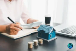Person calculating finances with a house model and coins on a desk, representing Rent Bank Loans.
